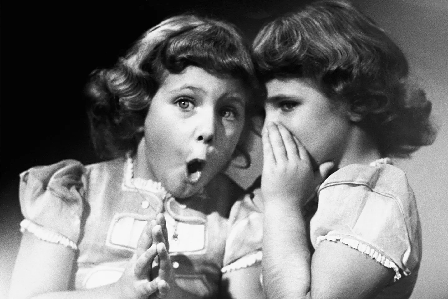 Black and white photograph of two young girls, one whispering a secret into the ear of the other who looks surprised